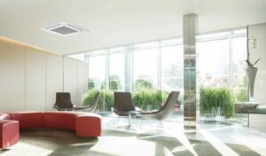 Modern lounge area with large windows, several black chairs, red curved seating, green plants by the windows, and a ceiling-mounted air conditioning unit. Natural light brightens the room.