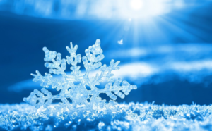 A close-up of a detailed snowflake resting on frosty ground, with sunlight shining in the blue-toned background and creating a bright, sparkling atmosphere.