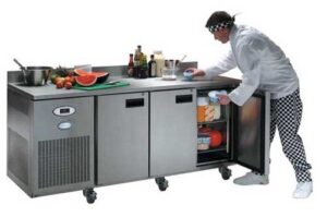 A chef in checkered pants cleans a stainless steel commercial prep table with storage, which holds ingredients and drinks; fruits and kitchen items are on the countertop.