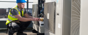 A worker wearing a blue hard hat and yellow safety vest kneels while servicing a rooftop air conditioning unit, using tools and inspecting the equipment outdoors.