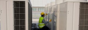 A worker in a yellow hard hat and safety vest performs maintenance on large HVAC units outdoors, kneeling between two rows of equipment on a rooftop.