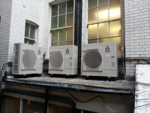 Three outdoor Mitsubishi air conditioning units are installed on a concrete platform attached to a white brick building, with multiple large windows and visible wiring in the background.
