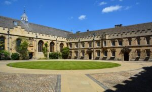 A historic stone building with arched windows surrounds a circular green lawn in a sunny courtyard, with cobblestone pathways and a bright blue sky above.