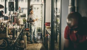A man stands by an open fridge in a bar, holding a bottle and looking up; another man leans forward in the foreground, partially out of focus. The bar counter is lined with bottles and equipment.