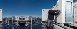 A technician in dark clothing works on a large air conditioning unit on a rooftop, surrounded by additional HVAC equipment under a clear blue sky.