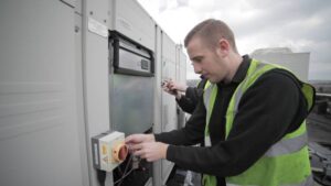 Air conditioning problems A technician in a high-visibility vest is working on electrical equipment outdoors, using tools to adjust a control box on a large industrial unit. Another person is partially visible in the background.