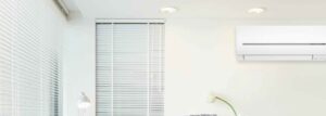 A minimalist room with white walls, a white window blind, a desk lamp, a flower in a vase, stacked books, and a white wall-mounted air conditioner.