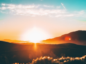Bright orange sun rises over rolling hills and mountains, casting a warm glow across the landscape. The sky is partly cloudy, with blue and golden hues blending above the silhouetted terrain.