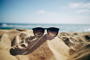 A pair of sunglasses sits on a sandy beach with the ocean and blue sky blurred in the background, evoking a relaxing summer vibe.