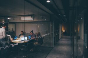 A group of people sit around a conference table in a glass-walled office, working on laptops and talking, while a hallway with dim lighting is visible through the glass wall.